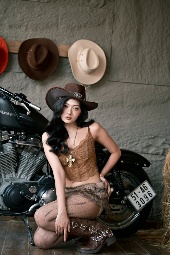 Home Trendy cowgirl in western attire posing with a motorcycle indoors.